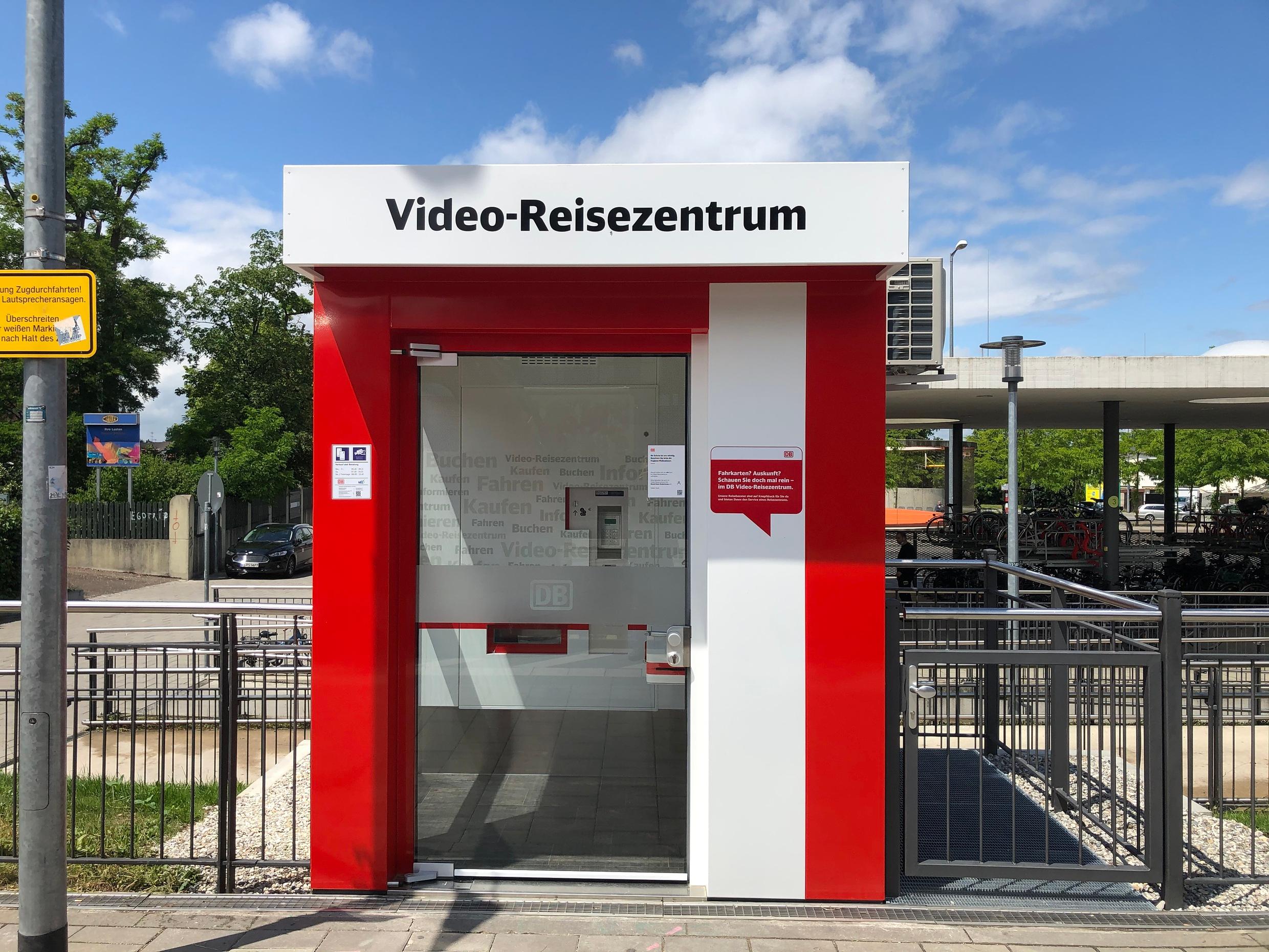 Eine rot-weiße Video-Reisezentrum-Kabine mit Glastür an einem Bahnsteig in München.