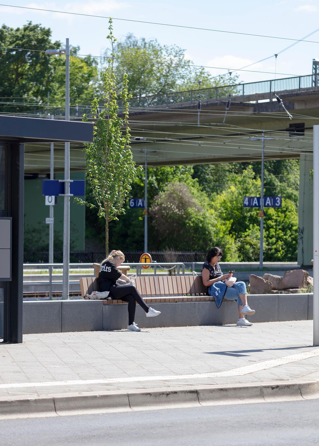 Zwei Personen sitzen auf den neuen Holzwartemöbeln am Vorplatz.