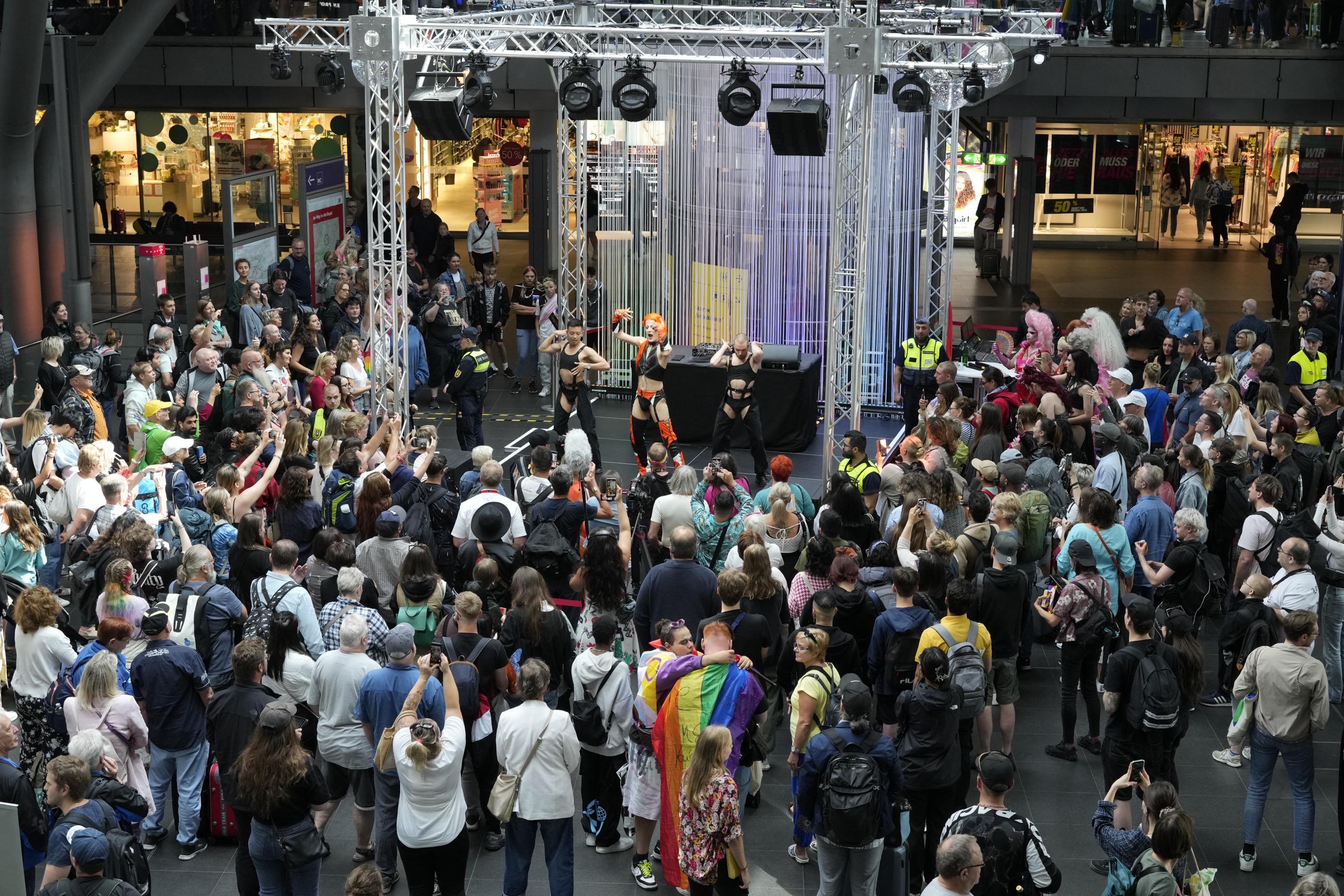 Drag queens dance and sing on the event stage at Berlin Central Station, surrounded by spectators.	